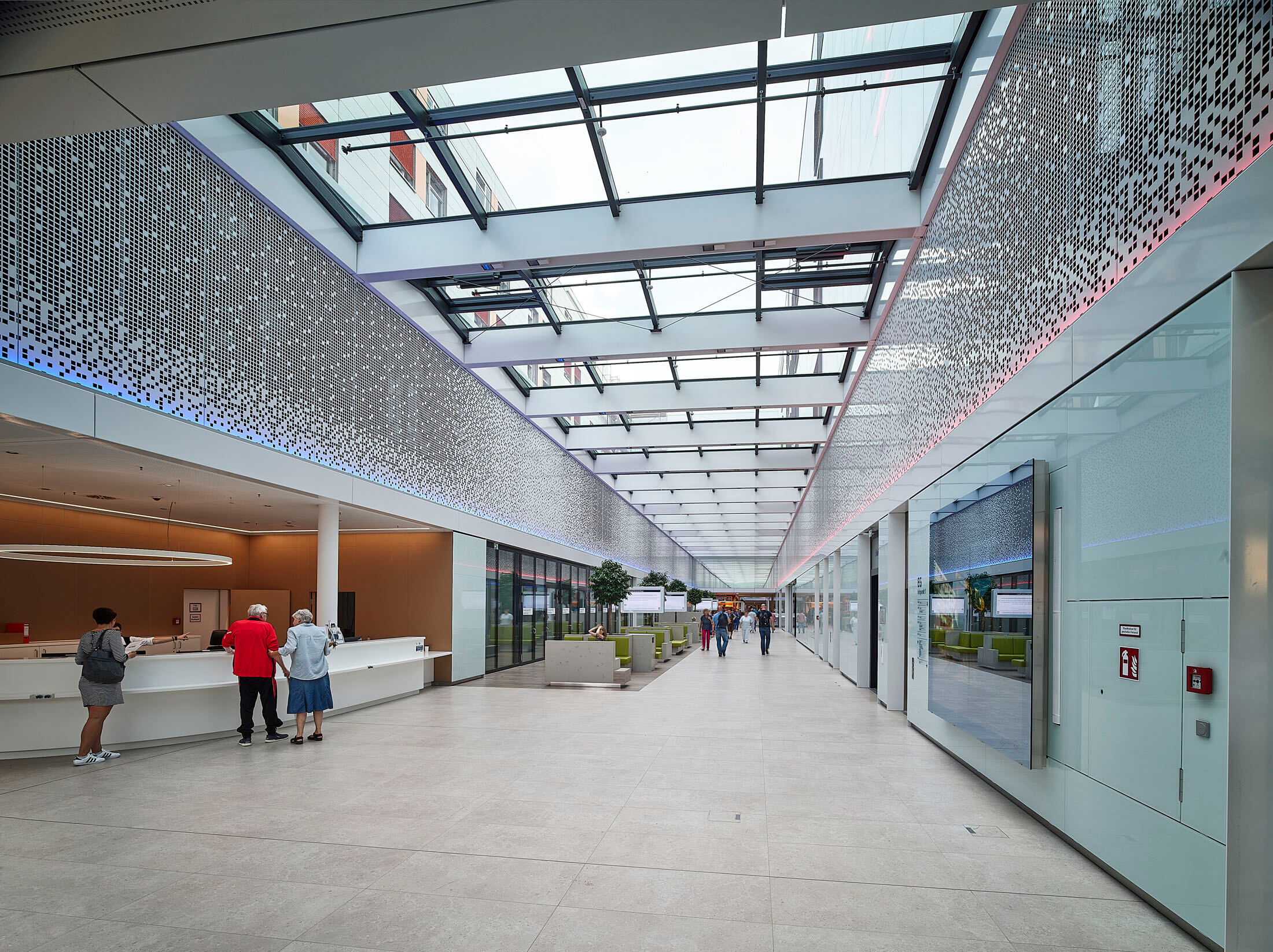 Große, moderne Eingangshalle mit verglastem Dach, LED-Beleuchtung, Empfangstresen und Besuchern, die durch die helle Passage gehen