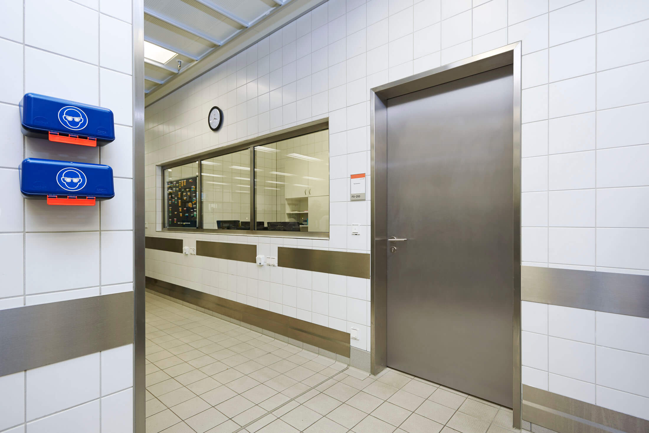 Hygienischer, gekachelter Betriebsflur mit Edelstahl-Tür, Wandfenster zum Arbeitsbereich und blauen Spendersystemen an der Wand