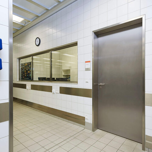 Hygienischer, gekachelter Betriebsflur mit Edelstahl-Tür, Wandfenster zum Arbeitsbereich und blauen Spendersystemen an der Wand