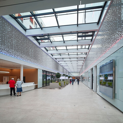 Große, moderne Eingangshalle mit verglastem Dach, LED-Beleuchtung, Empfangstresen und Besuchern, die durch die helle Passage gehen